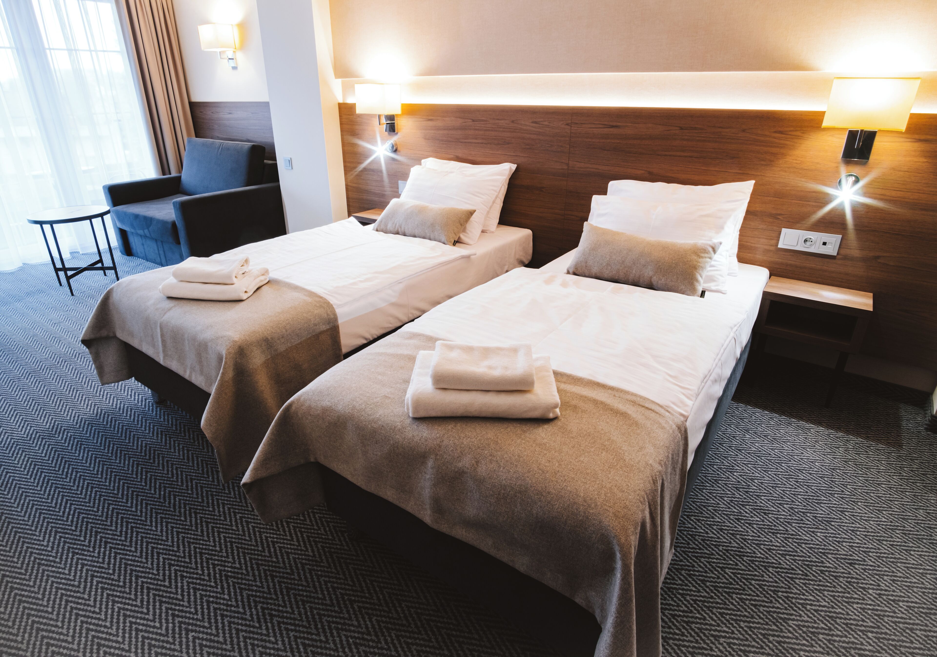 A hotel room with two beds, a desk, a chair, and a lamp.
