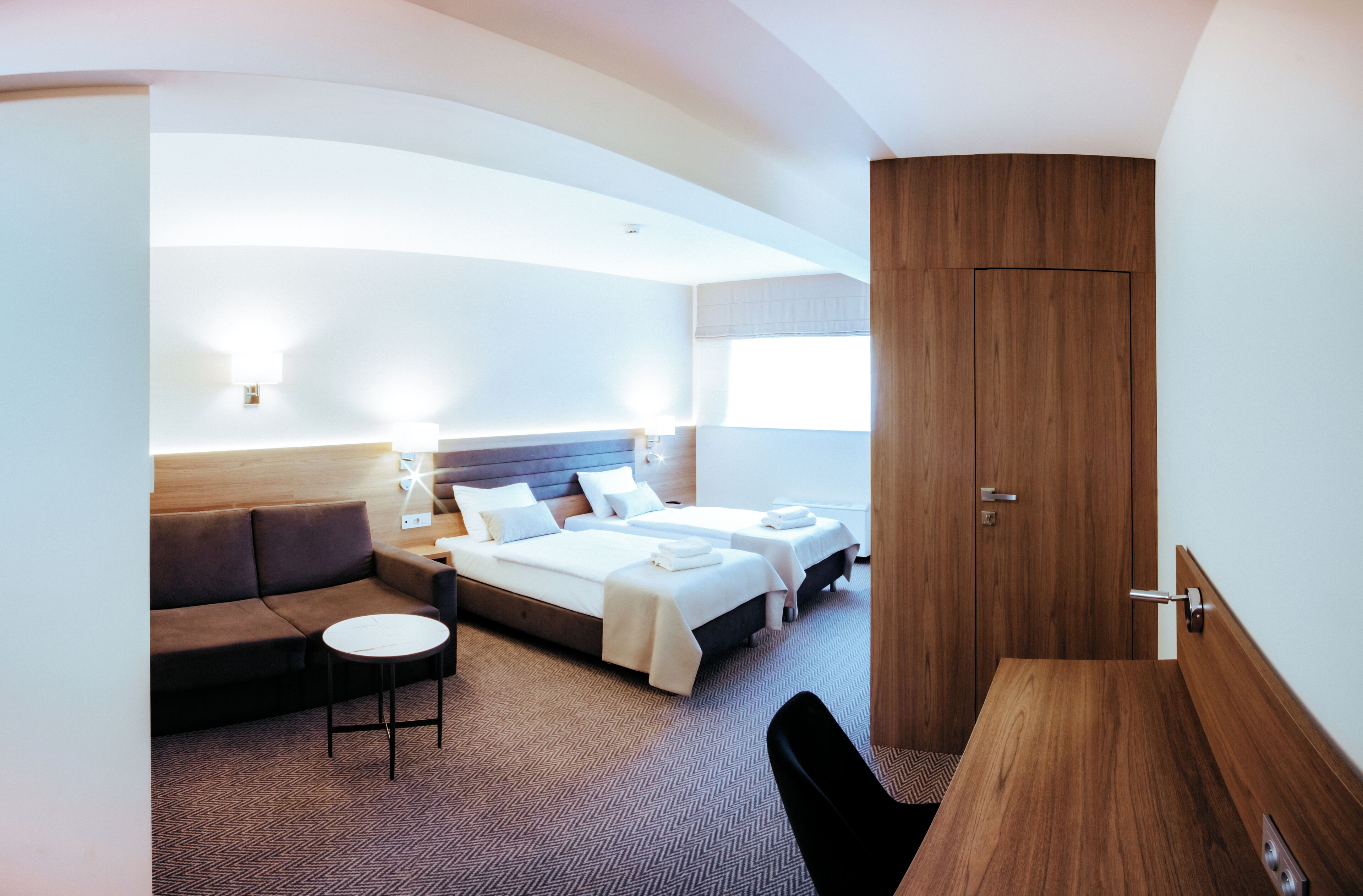 Quarto de hotel moderno com uma cama grande, um sofá, uma mesa redonda pequena e um guarda-roupa de madeira.