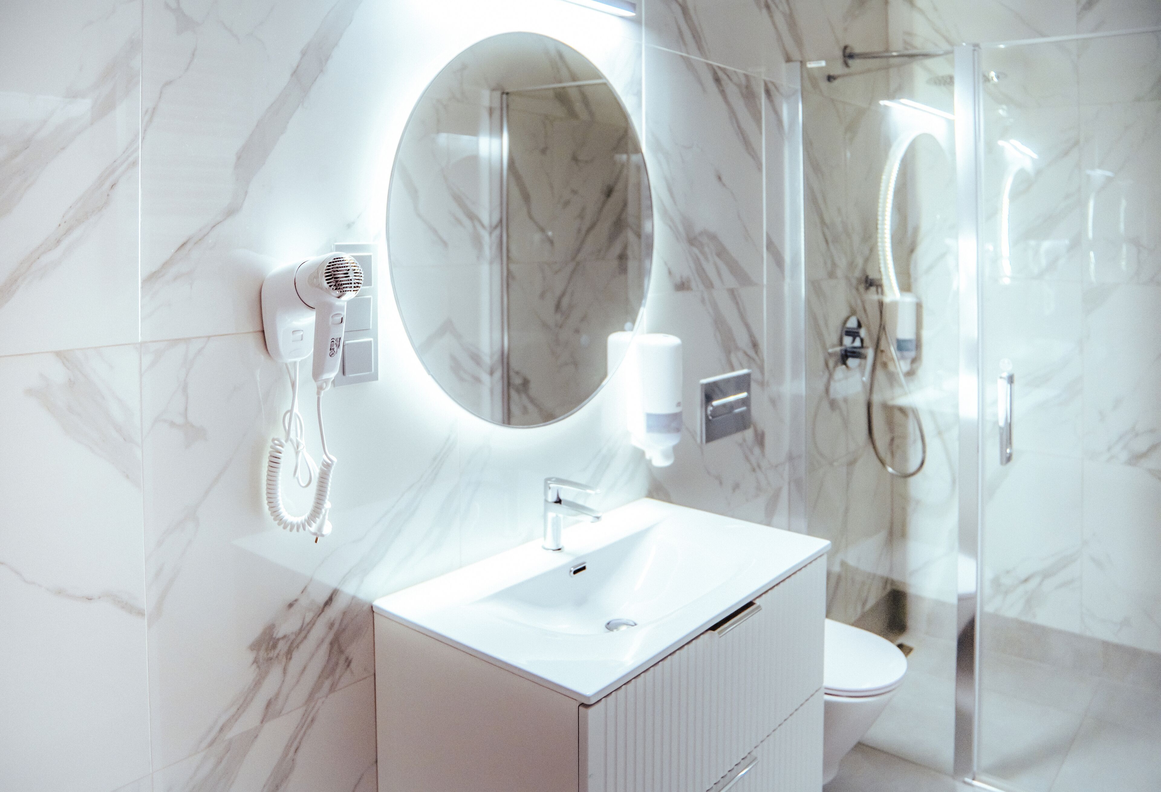 A modern bathroom with a white sink, a round mirror, a handheld hair dryer, and a glass-enclosed shower.