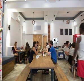 A modern dining area with wooden tables and chairs, a high ceiling with pendant lights, and a visible kitchen area in the background.