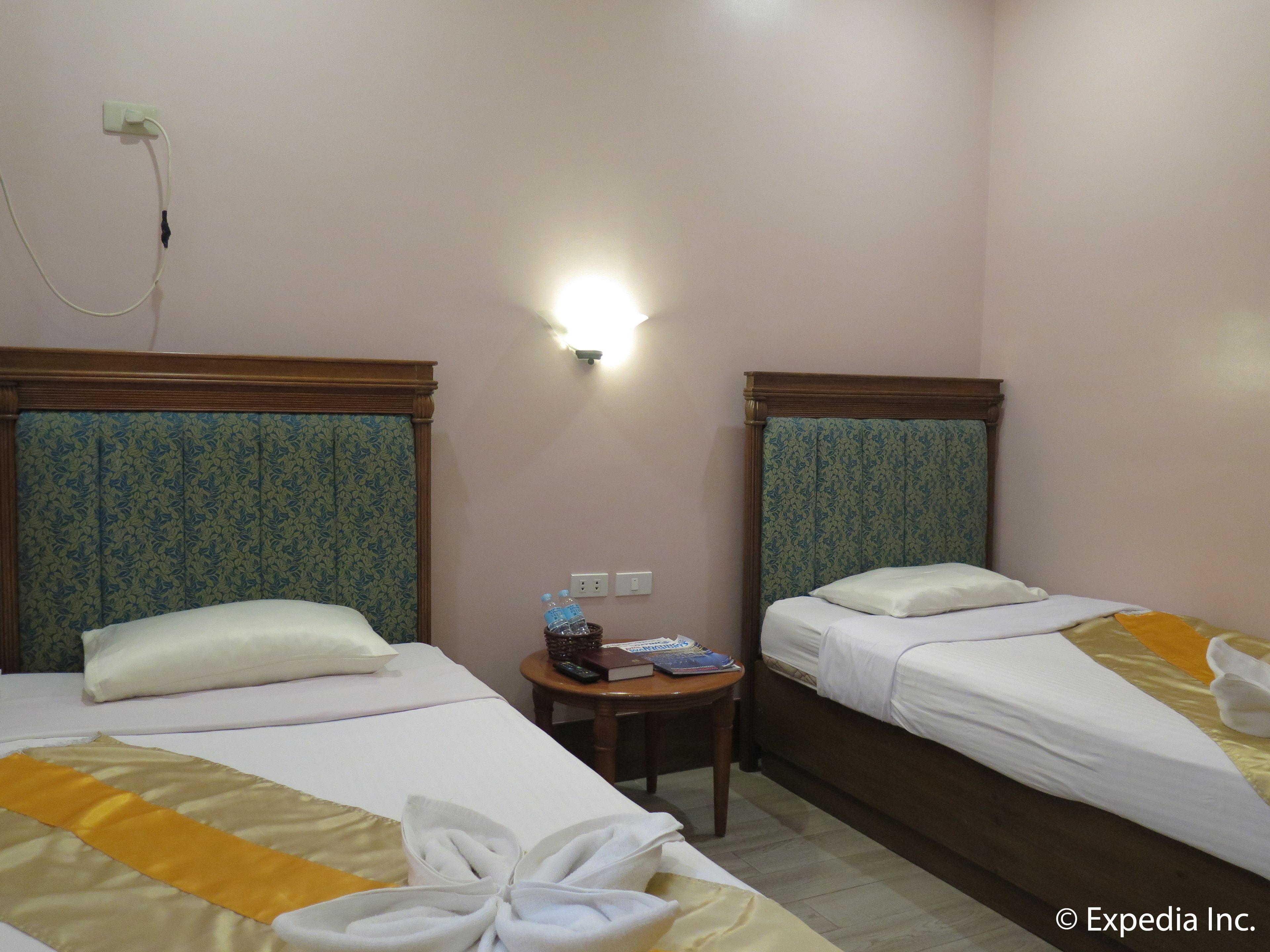 A hotel room with two beds, each with a headboard, a bedside table with a remote control, books, and a bottle of water.