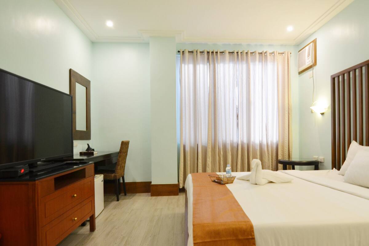 A hotel room with a bed, a desk with a television, a chair, and a window with curtains.