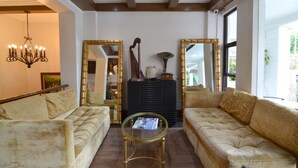 Un salon spacieux avec deux canapés de couleur crème, une table basse en verre et un lustre. De grands miroirs ornent les murs, et un meuble noir abrite l’équipement audiovisuel.