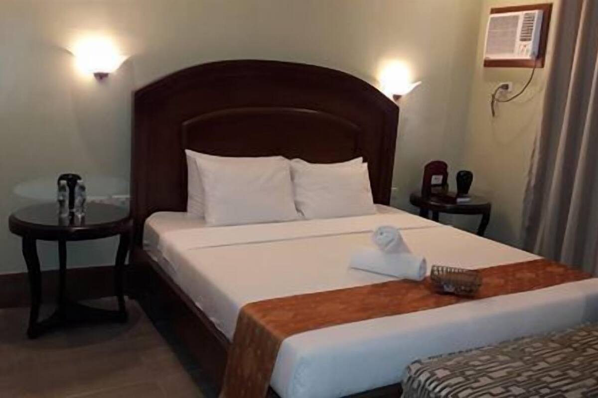 A hotel room with a wooden headboard, a bed with white linens, a small table with a glass top, a chair, and a wall-mounted air conditioning unit.