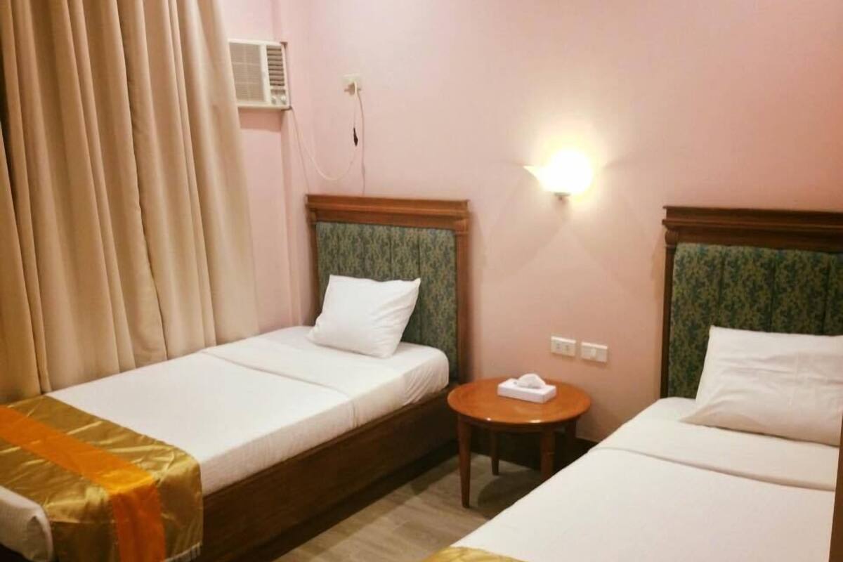 A hotel room with two single beds, a small round table, a wall-mounted air conditioner, and a light fixture.