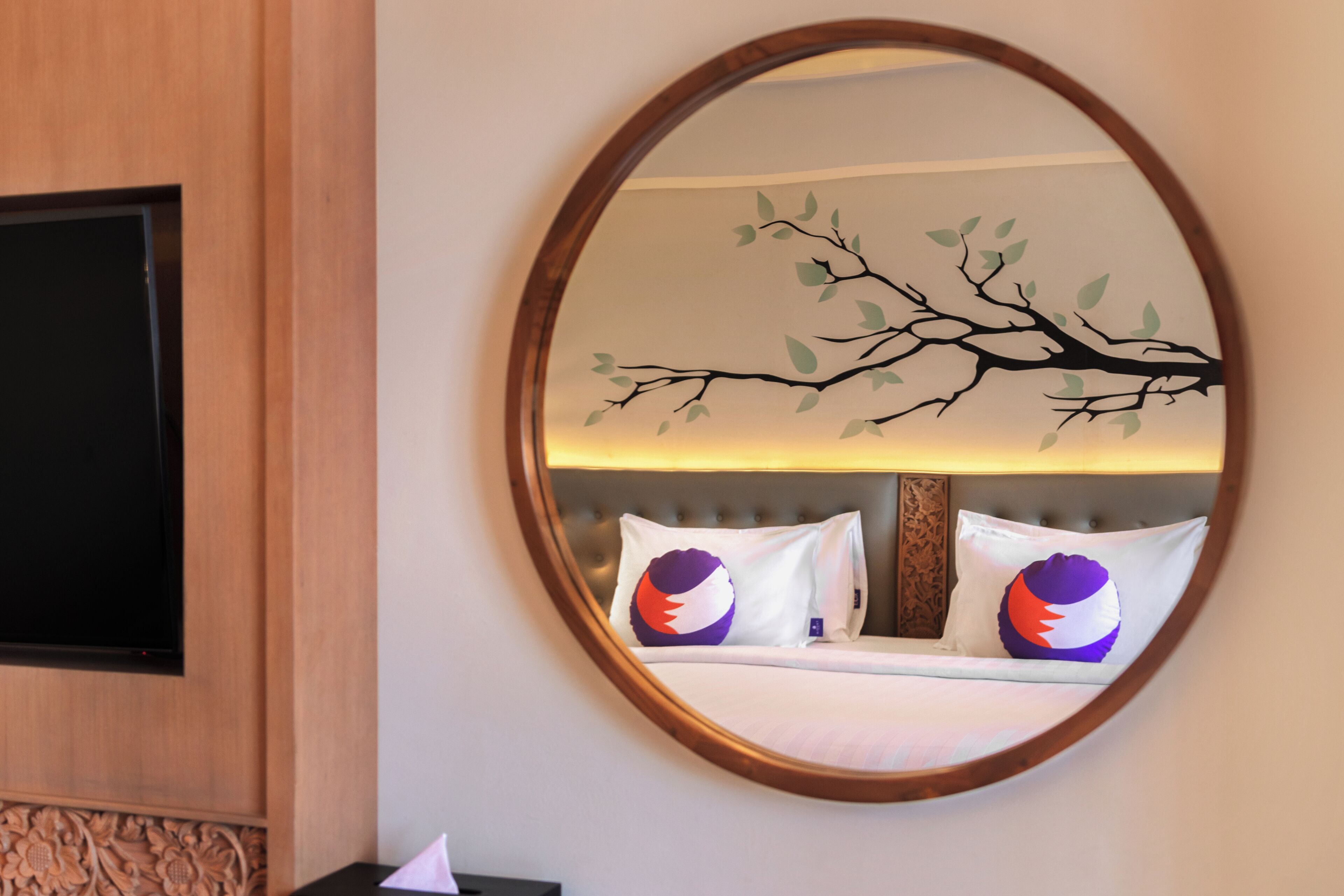 A hotel room with a large round mirror, a flat-screen TV, a bed with white bedding, and a decorative wall mural.