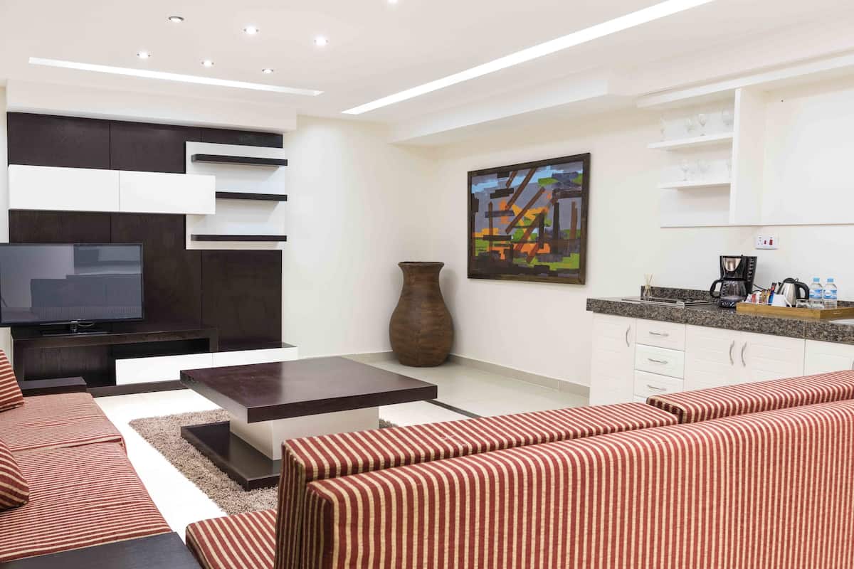 A modern living room with a sofa set, a coffee table, a TV, a vase, and a kitchen area with cabinets and a coffee machine.