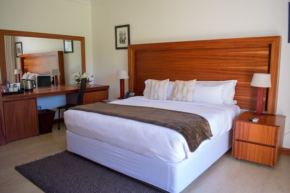 Une chambre d’hôtel avec un grand lit, une tête de lit en bois, des tables de chevet avec des lampes, un bureau avec une machine à café et un miroir.