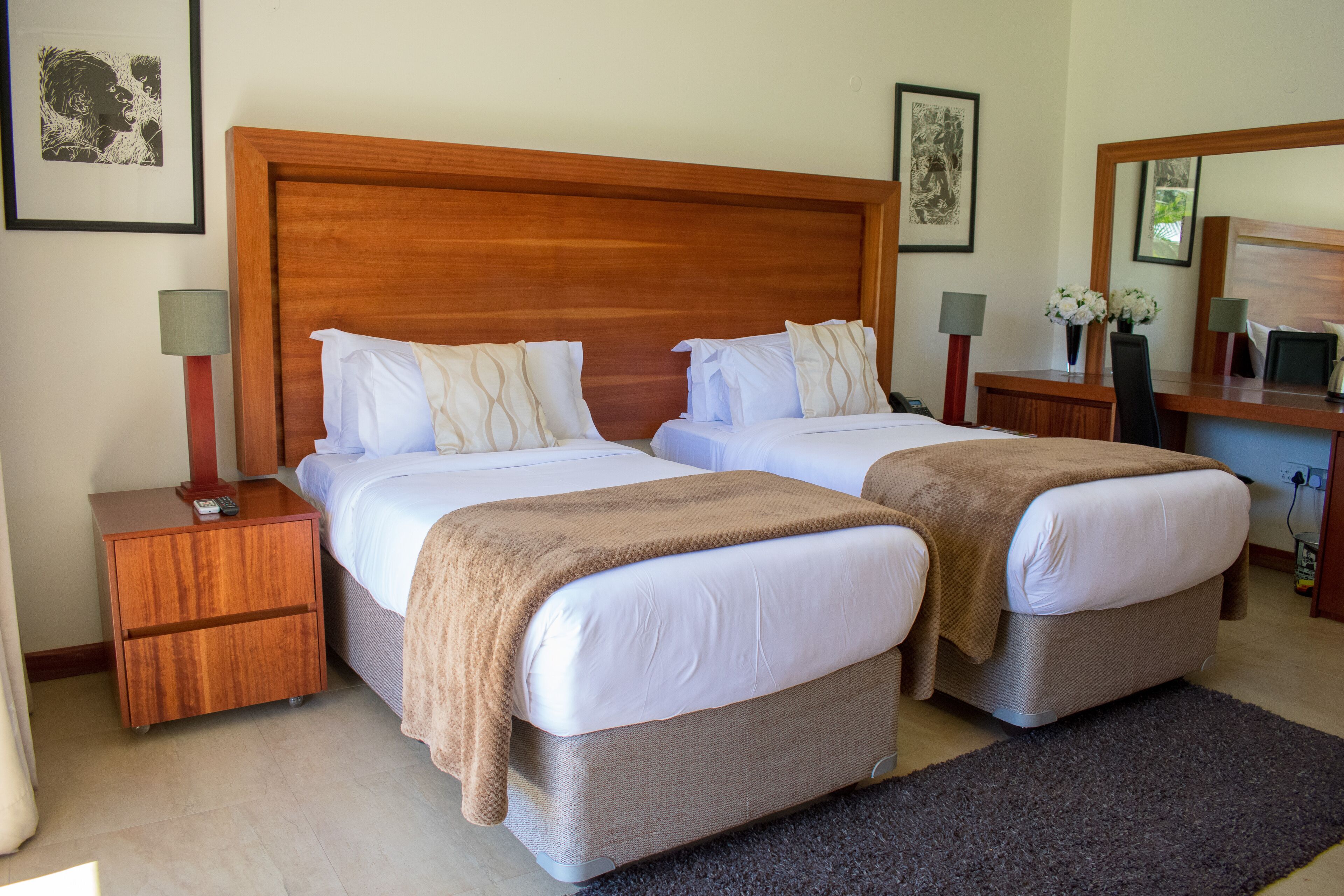 Una cama doble con cabecera de madera, dos mesitas de noche con lámparas, un escritorio con espejo y un florero con flores.