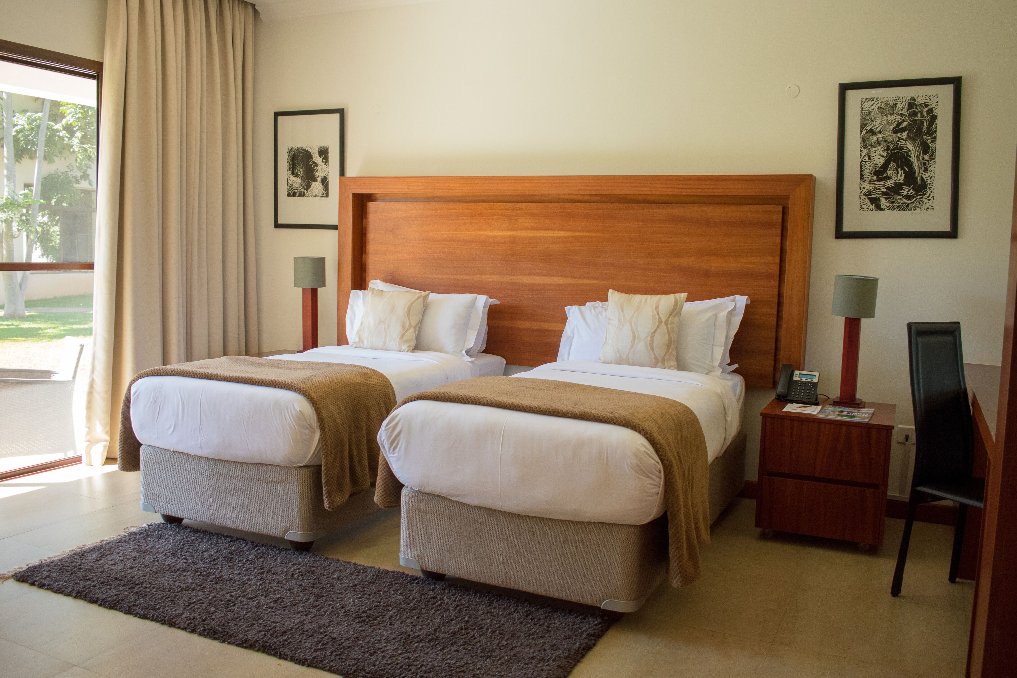 A hotel room with two beds, a wooden headboard, bedside lamps, a small wooden nightstand, and a view of the outdoors.