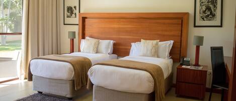 A hotel room with two beds, a wooden headboard, bedside lamps, a small wooden nightstand, and a view of the outdoors.