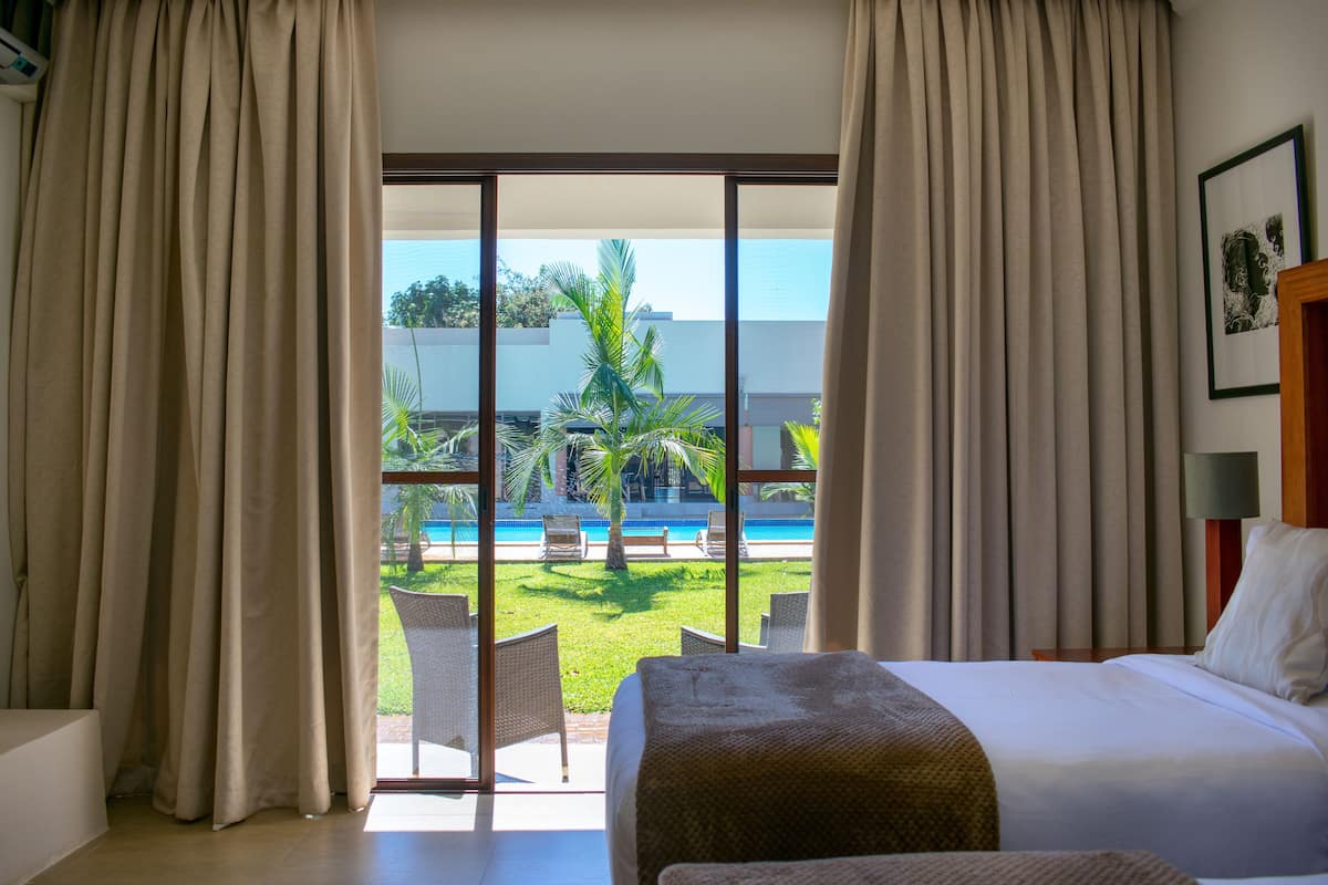 A hotel room with a bed, a view of a pool, and a balcony with chairs.