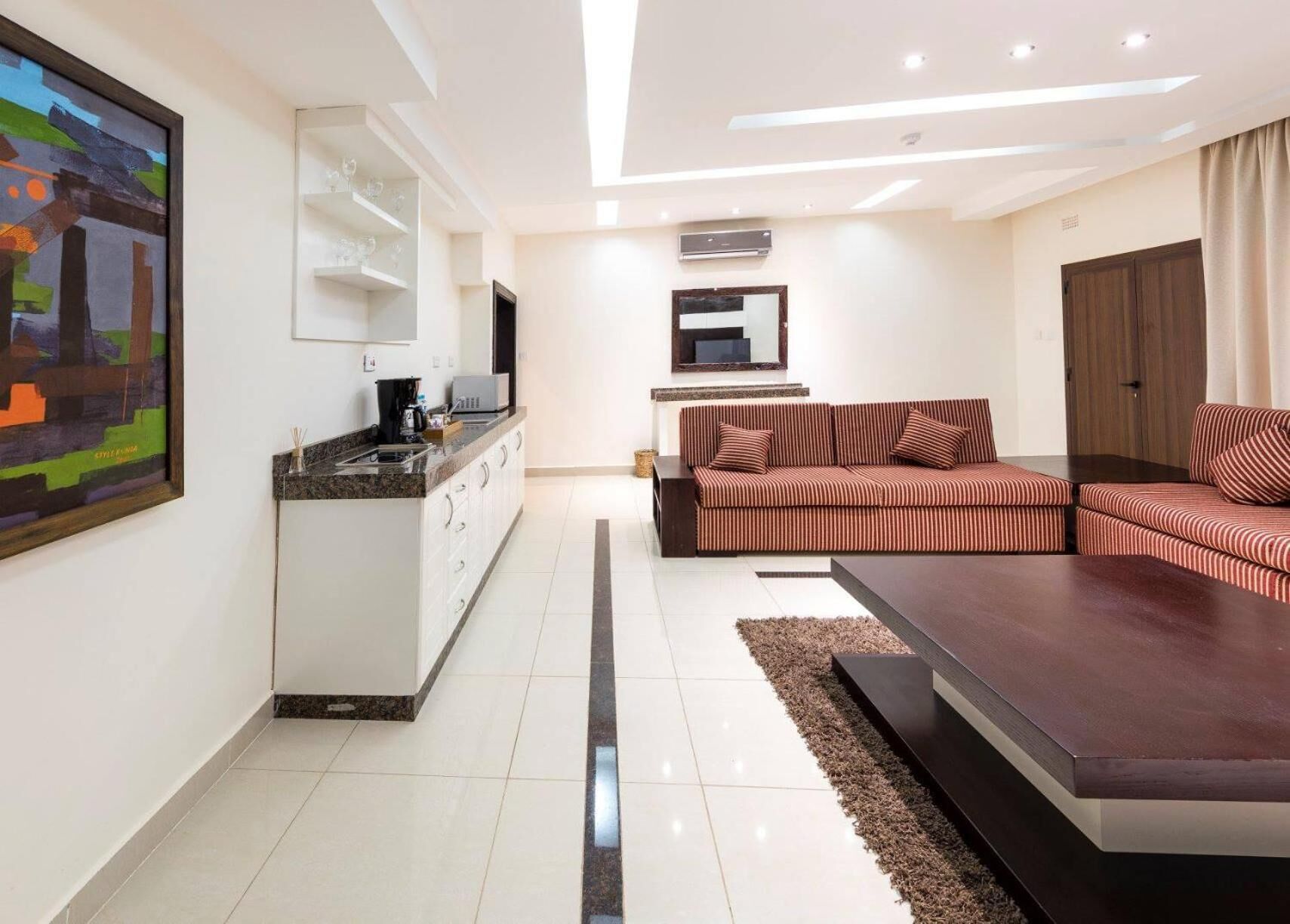 A modern living room with a sofa, a coffee table, a kitchen area with a sink and cabinets, and a large painting on the wall.