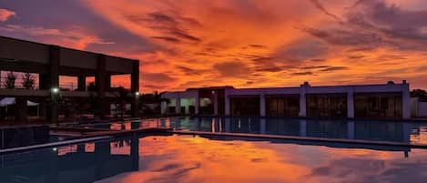 A swimming pool with a sunset sky reflected on the water, surrounded by buildings.