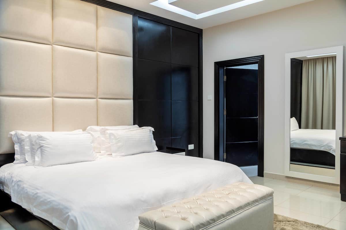 A modern bedroom with a large bed, a tufted headboard, a dark wardrobe, and a mirror reflecting the bed.