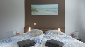 A neatly made bed with striped bedding, a painting of a beach scene above it, and a nightstand with a lamp and a pink flower.