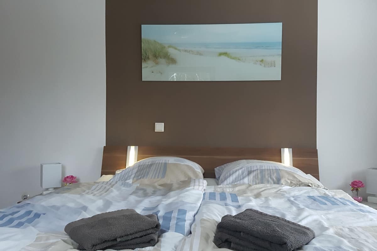 Un lit bien fait, avec une literie à rayures, un tableau représentant une scène de plage au-dessus, et une table de chevet avec une lampe et une fleur rose.