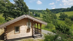 A small wooden cabin with a porch, surrounded by a lush green landscape and hills.