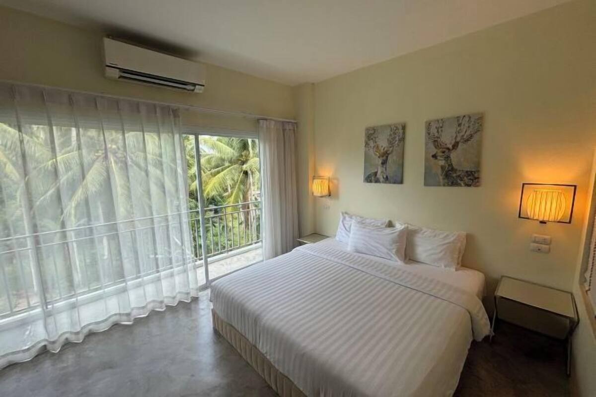 A hotel room with a large bed, a balcony with curtains, two wall-mounted lamps, and artwork on the walls.