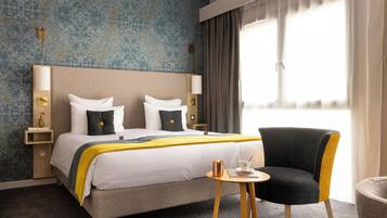 A hotel room with a large bed, two bedside tables, a chair, a small table, and a window with curtains.