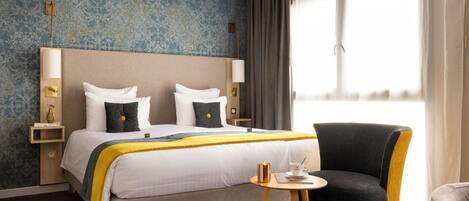 Habitación de hotel con una cama grande, dos mesitas de noche, una silla, una mesita pequeña y una ventana con cortinas.