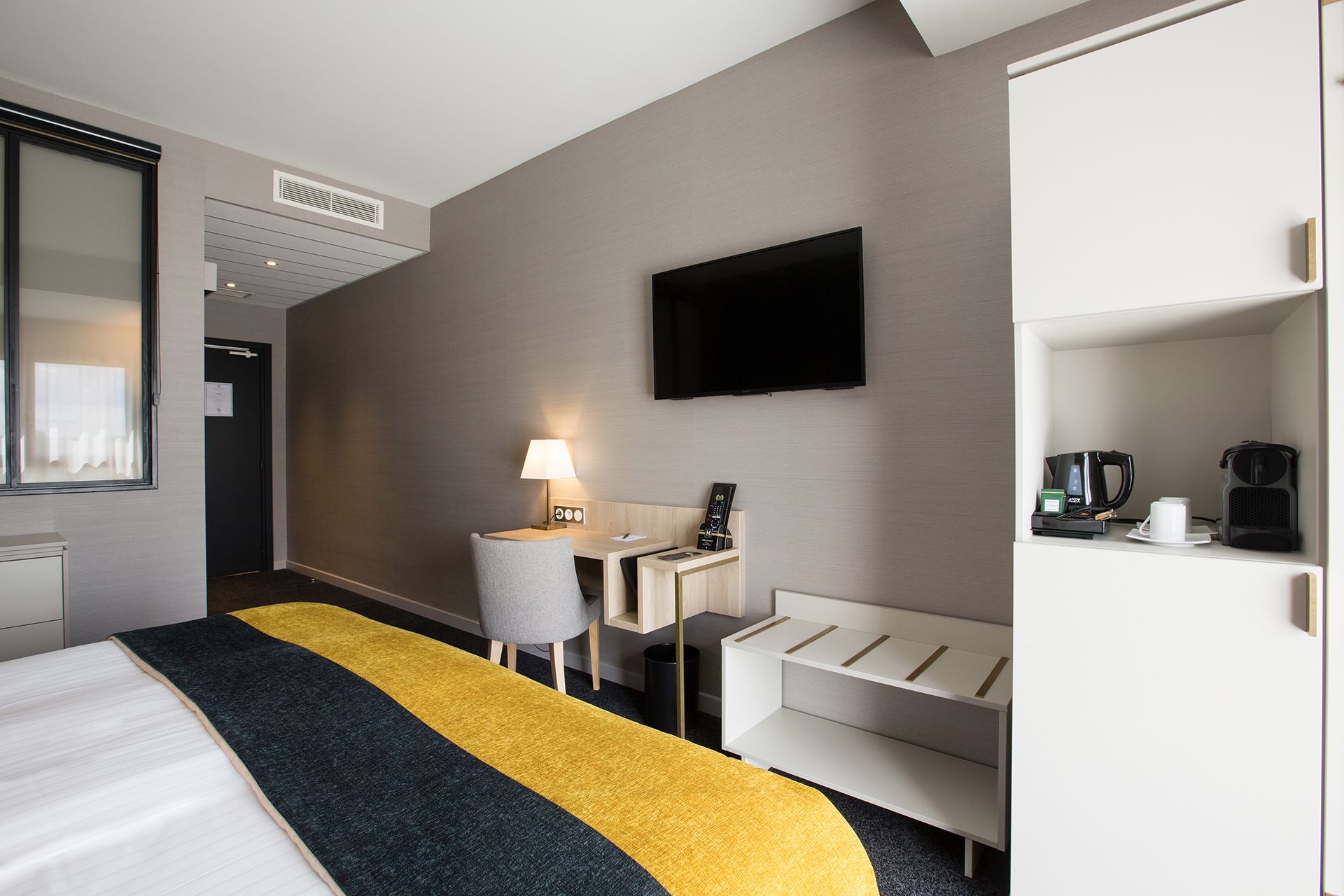 Une chambre d’hôtel équipée d’un lit, d’un bureau avec une lampe, d’une télévision, d’une machine à café et d’un mini-bar.