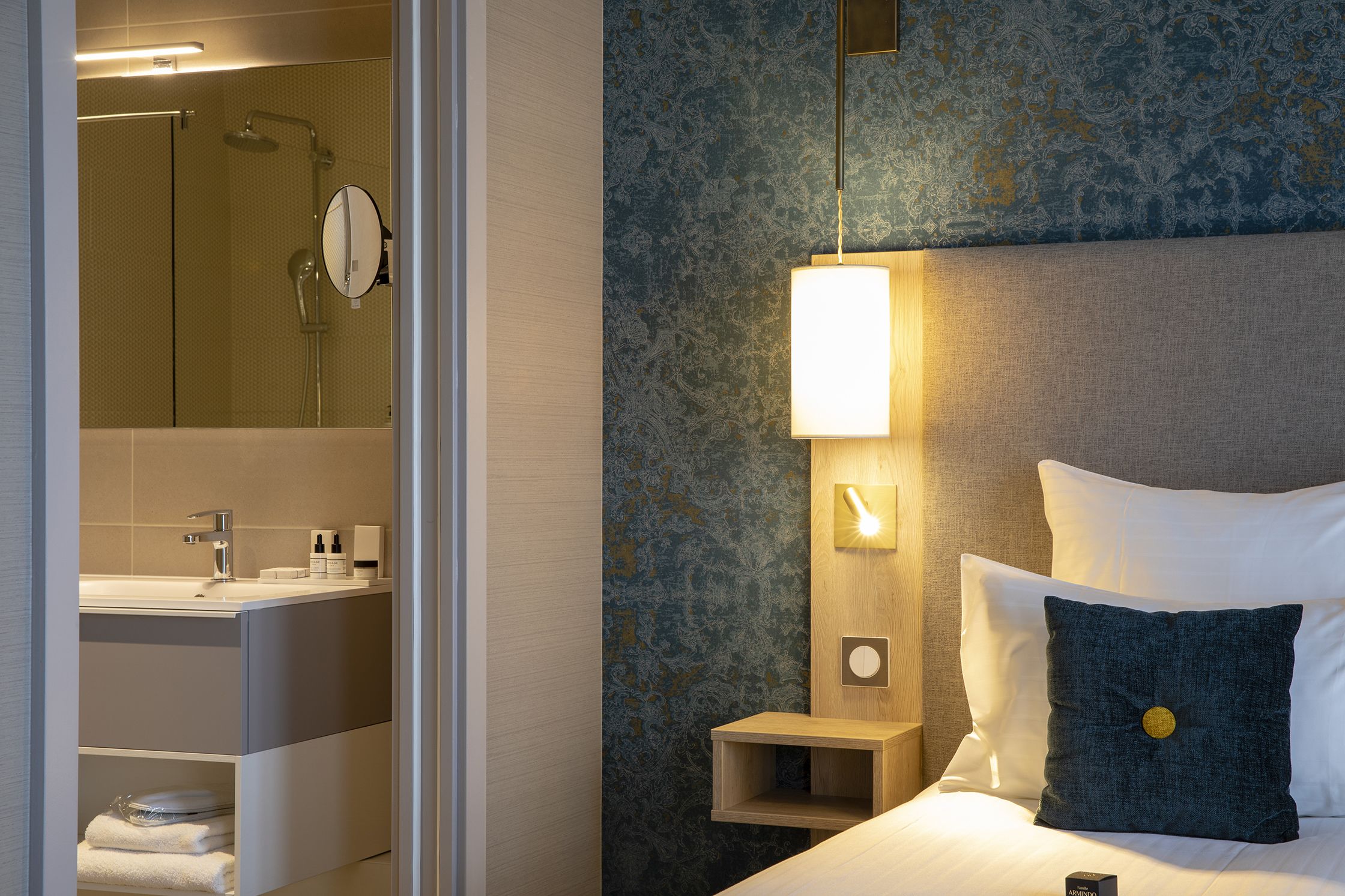 A modern hotel room with a bathroom, a bedside table with a lamp, a bed with pillows, and a nightstand.