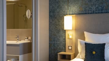 A modern hotel room with a bathroom, a bedside table with a lamp, a bed with pillows, and a nightstand.