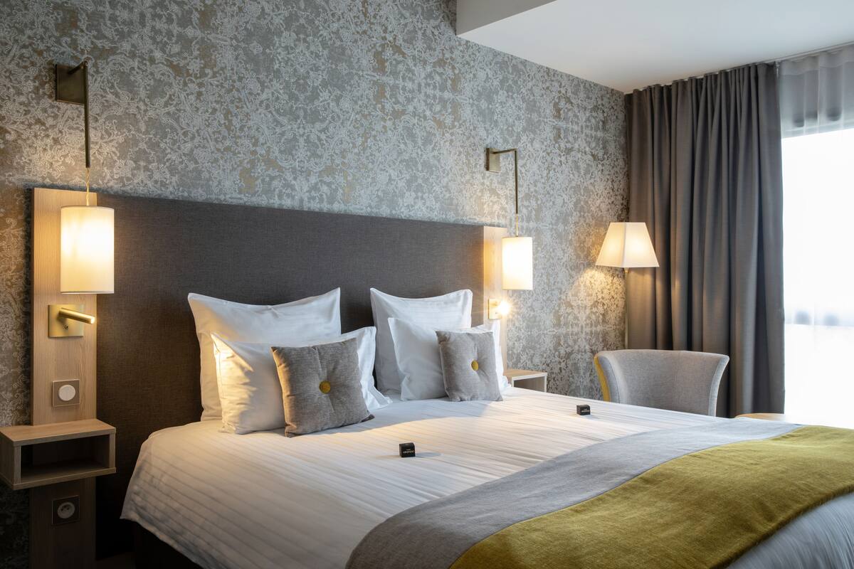 A hotel room with a large bed, two bedside tables, a nightstand, a chair, and a window with curtains.