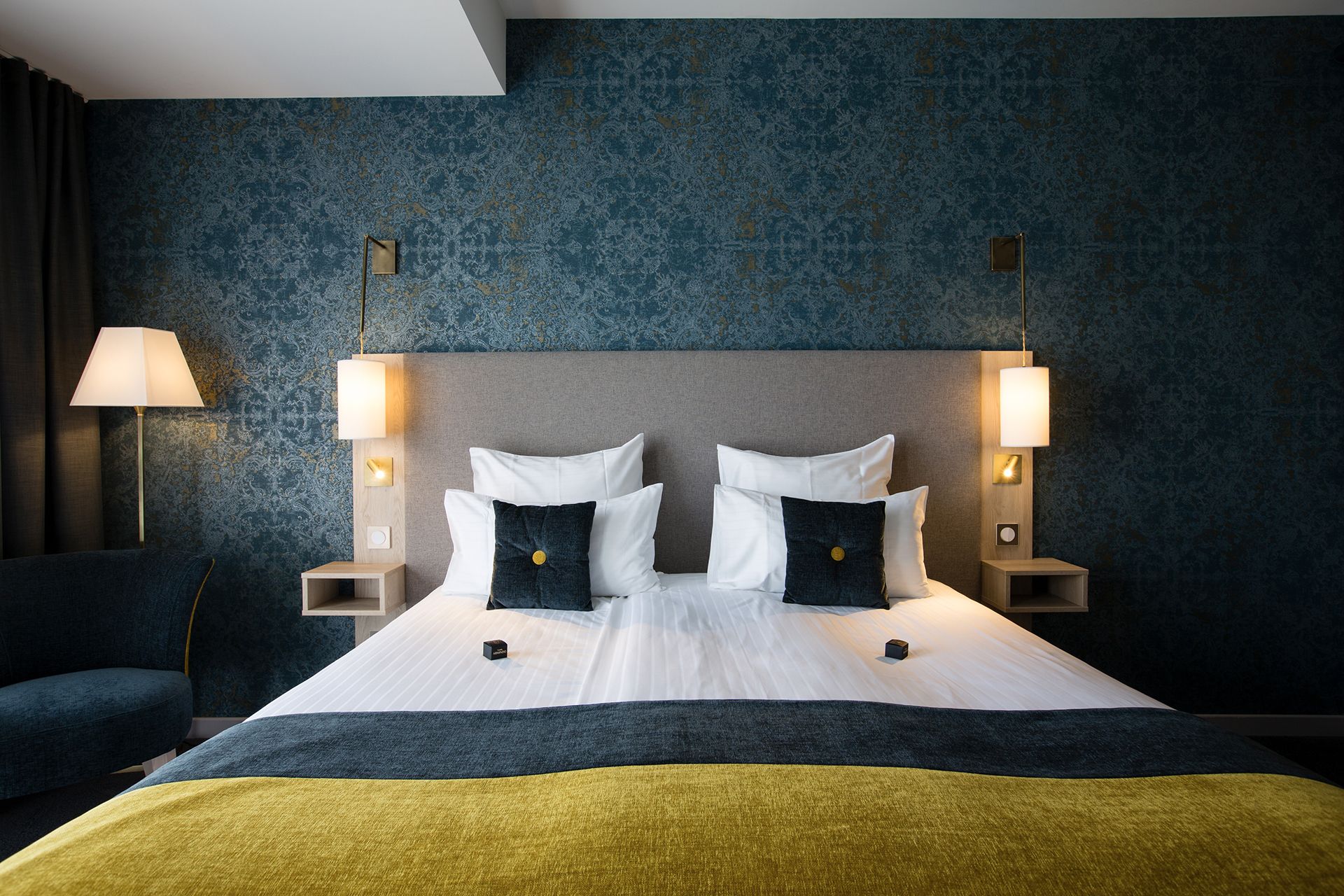 Une chambre d’hôtel avec un grand lit, deux lampes de chevet, une tête de lit et un mur de couleur bleu foncé orné d’un motif.