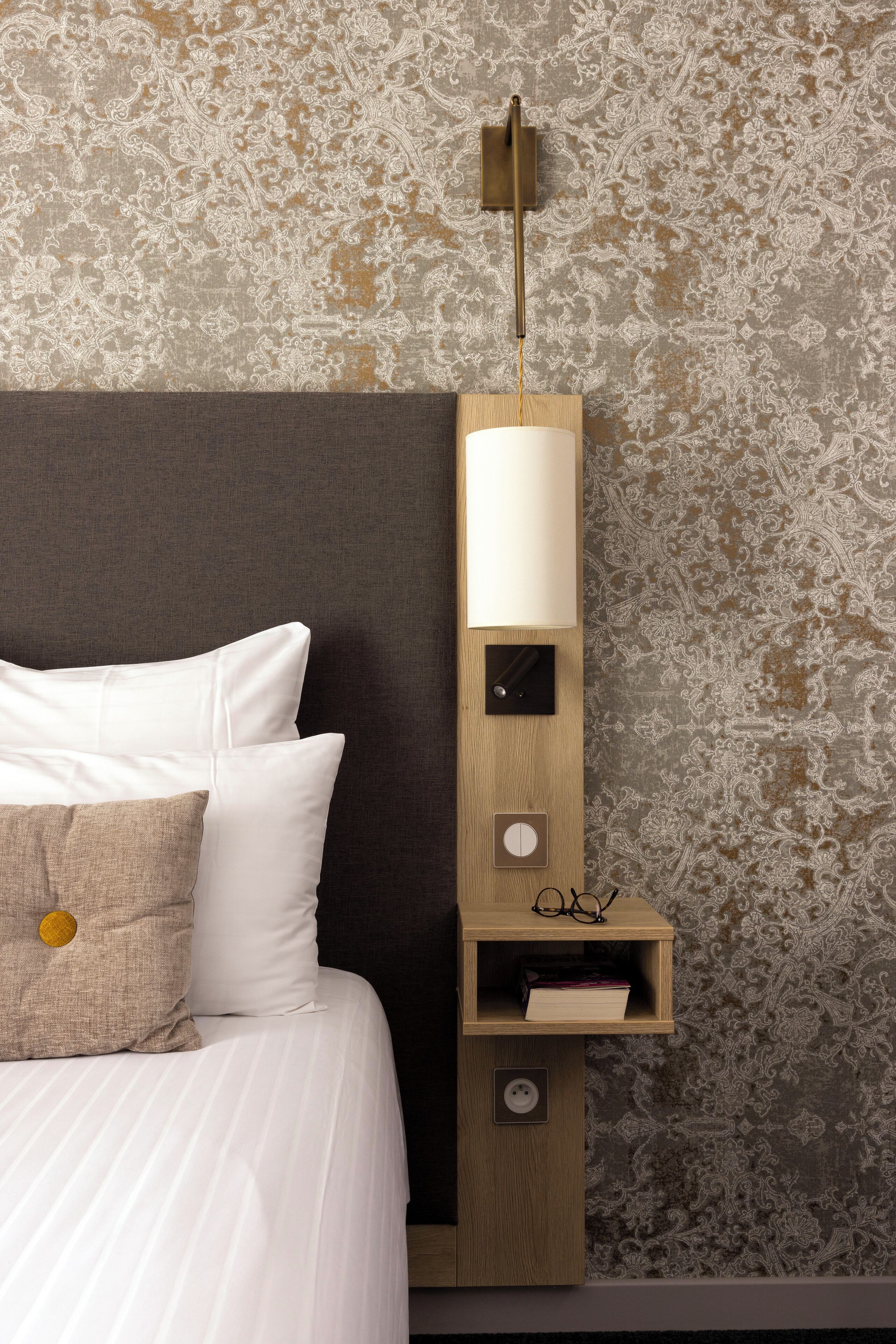 Un lit bien fait, avec du linge de lit blanc et un oreiller beige, une table de chevet en bois avec une lampe, et un mur recouvert d’un papier peint à motifs.