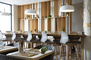 A modern dining area with wooden tables and chairs, pendant lighting, and plants on shelves. - James Boutique Hôtel (Colmar)