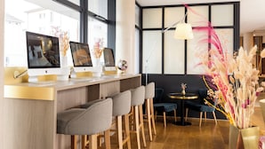 A modern bar area with Apple iMacs on a counter, bar stools, a round table with a lamp, and decorative plants. - James Boutique Hôtel (Colmar)
