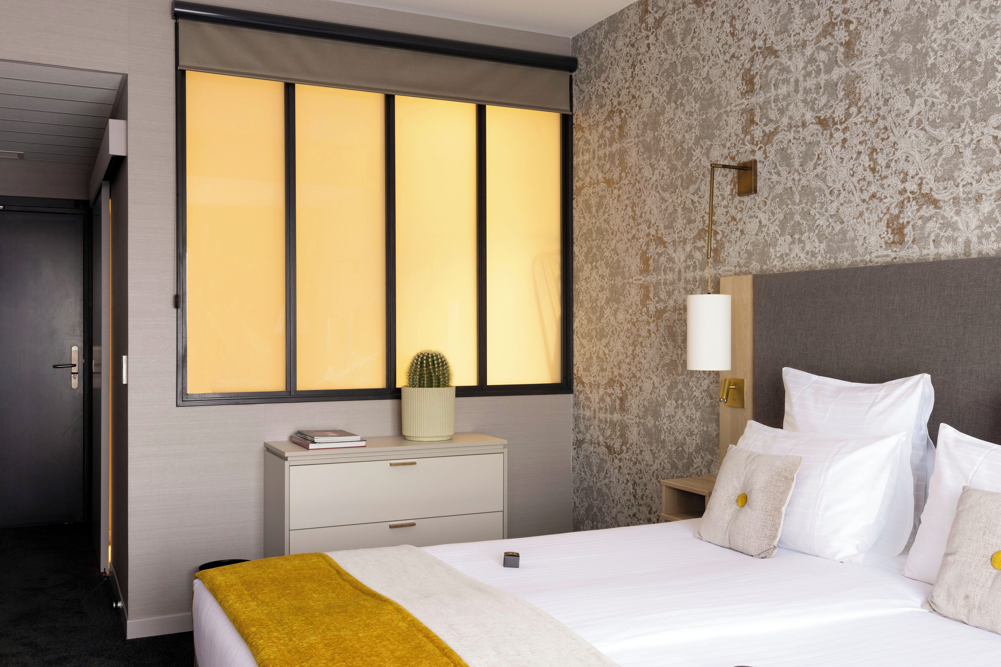 A hotel room with a bed, a nightstand, a lamp, and a window with curtains.