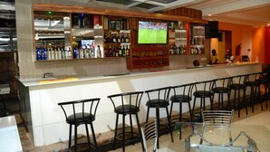 A bar area with a long counter, multiple stools, and a television screen displaying a sports event. - La Posh Hotel (Kigali)