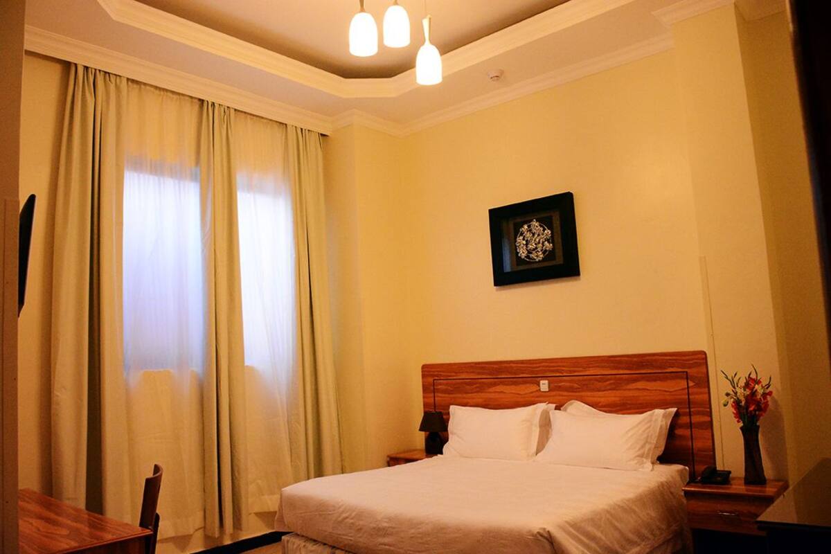 A hotel room with a bed, a nightstand, a lamp, a framed picture on the wall, and a window with curtains.