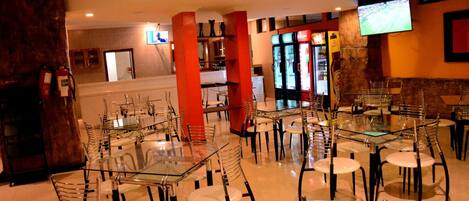 Un restaurante con mesas y sillas de vidrio, pilares rojos y un televisor montado en la pared.