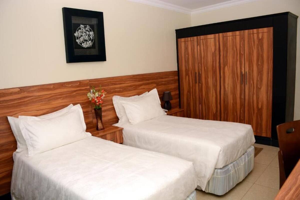 A hotel room with two beds, a wooden wardrobe, a framed picture on the wall, and a small table with a vase of flowers.