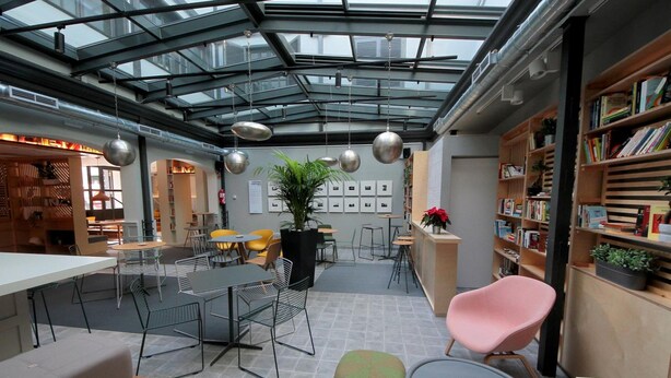 A modern indoor space with a glass ceiling, tables, chairs, and a bookshelf. - Complutum Hostel (Alcala de Henares)