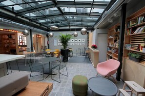 A modern indoor space with a glass ceiling, tables, chairs, and a bookshelf. - Complutum Hostel (Alcala de Henares)