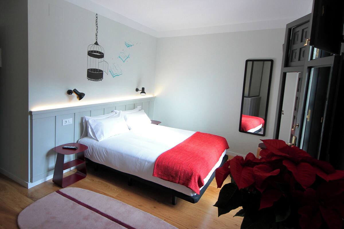 Un dormitorio con cama, mesita de noche, lámpara, espejo y una planta grande en maceta.