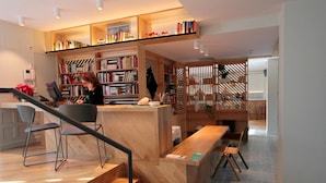 A modern library with wooden bookshelves, a reception desk, and a staircase. - Complutum Hostel (Alcala de Henares)