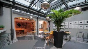 A modern indoor space with tables and chairs, a large potted plant, and a wall with framed pictures. - Complutum Hostel (Alcala de Henares)