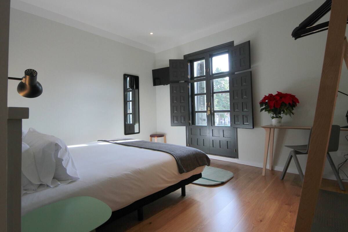 Un dormitorio con una cama, un escritorio, una silla, una planta en maceta y una ventana con contraventanas.