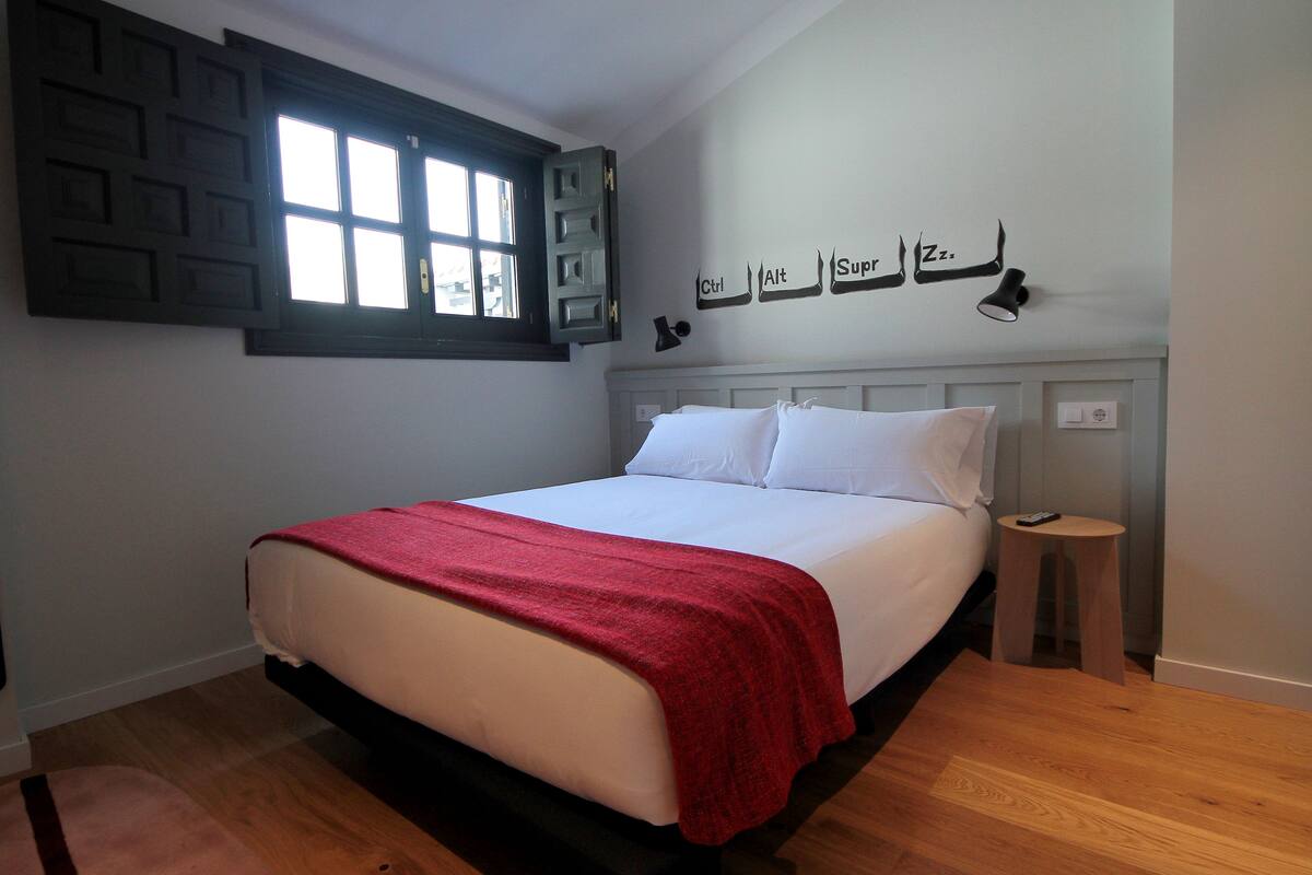Una cama bien hecha con una colcha roja, una mesita de noche con una lámpara y una ventana con un motivo geométrico en la pared.