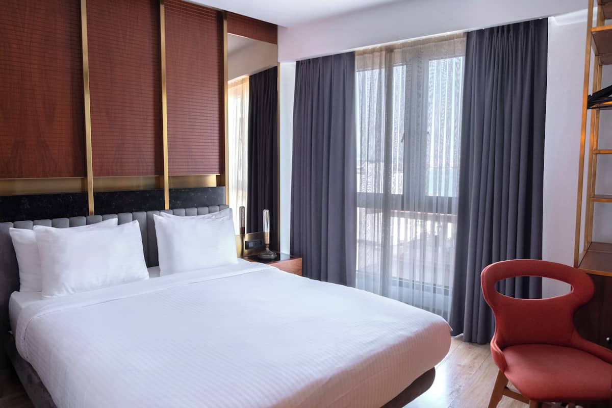 Une chambre d’hôtel avec un grand lit, un fauteuil rouge, une table de chevet et une fenêtre avec des rideaux.