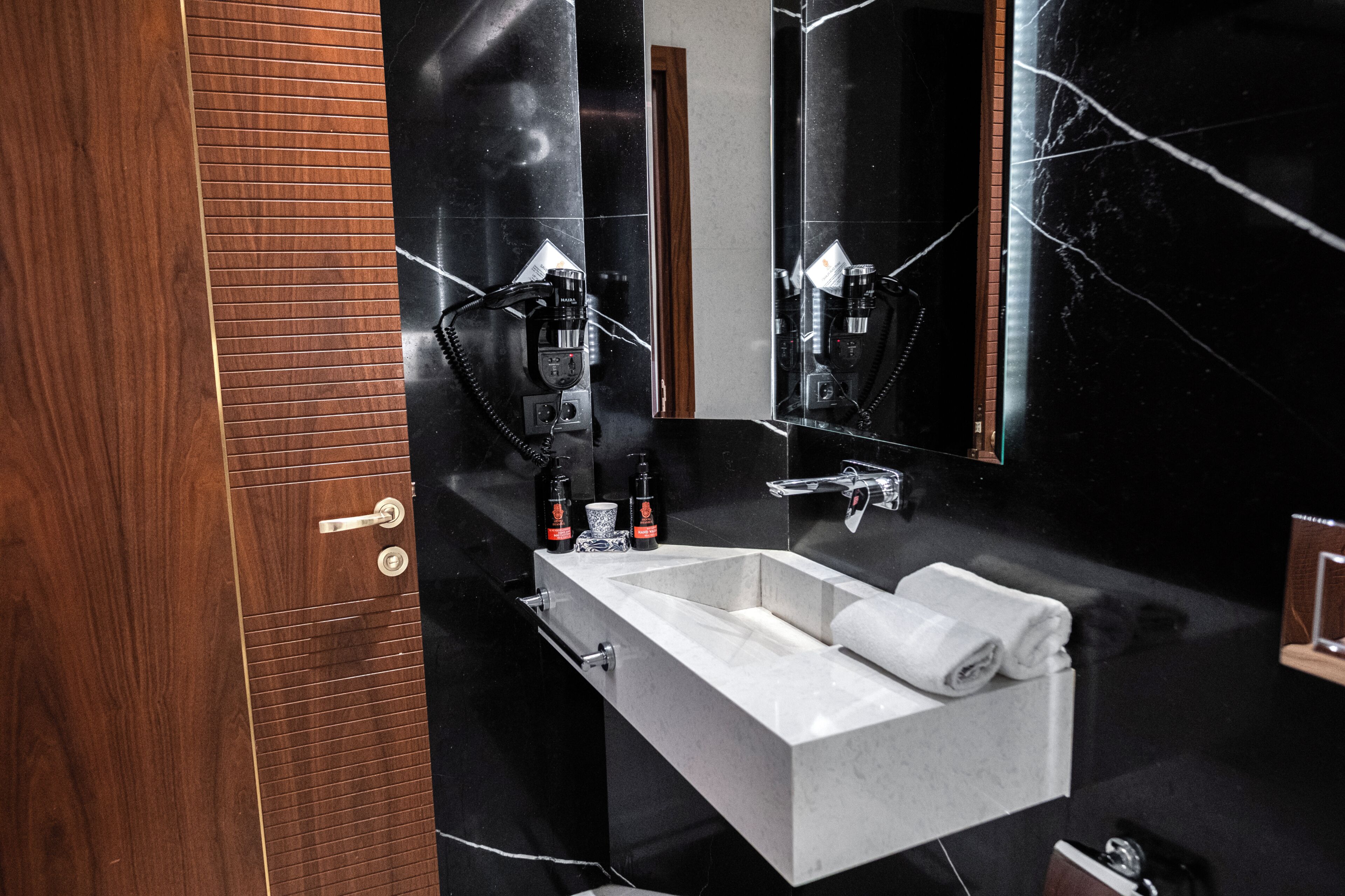 Une salle de bain moderne avec un lavabo blanc, des murs en marbre noir et un grand miroir.