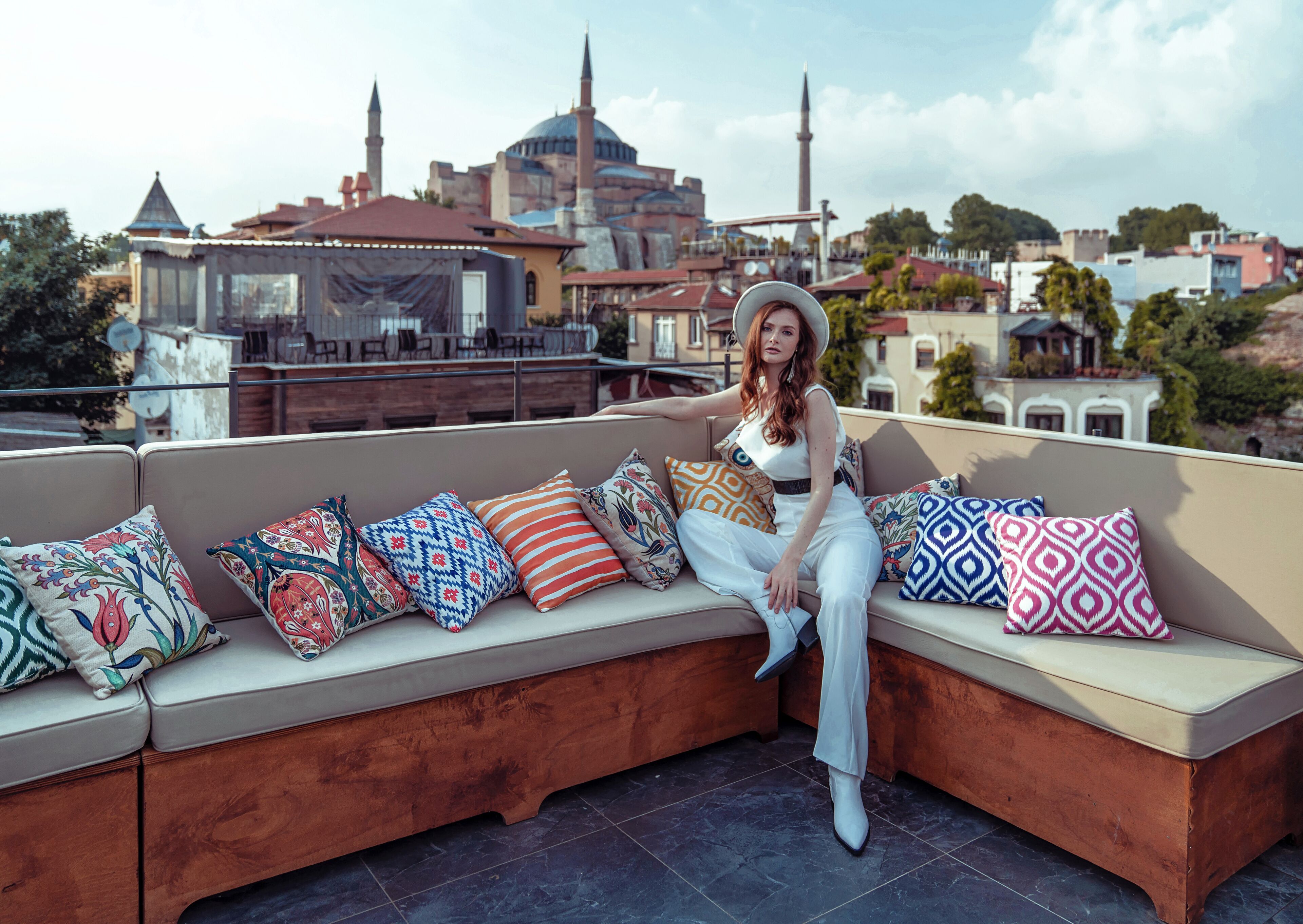 A woman sitting on a curved sofa with colorful cushions, overlooking a cityscape with historic buildings and a mosque.