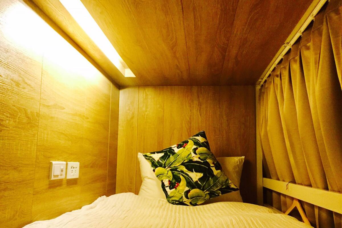 A room with wooden walls, a bed with a white bedspread, a pillow with a tropical leaf pattern, and a curtain.