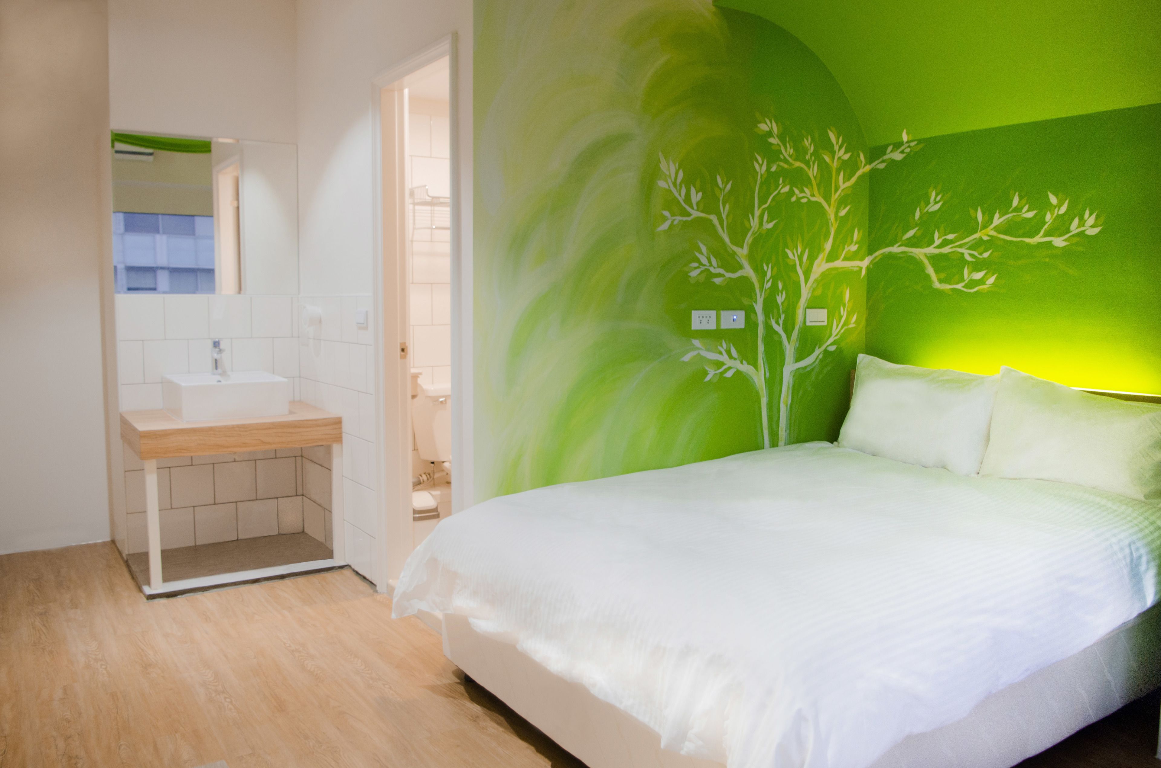 A bedroom with a bed, white bedding, a green wall mural, and a bathroom with a sink and mirror.